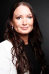 Emily Dean is facing the camera. She has long dark hair and is wearing a white jacket. 